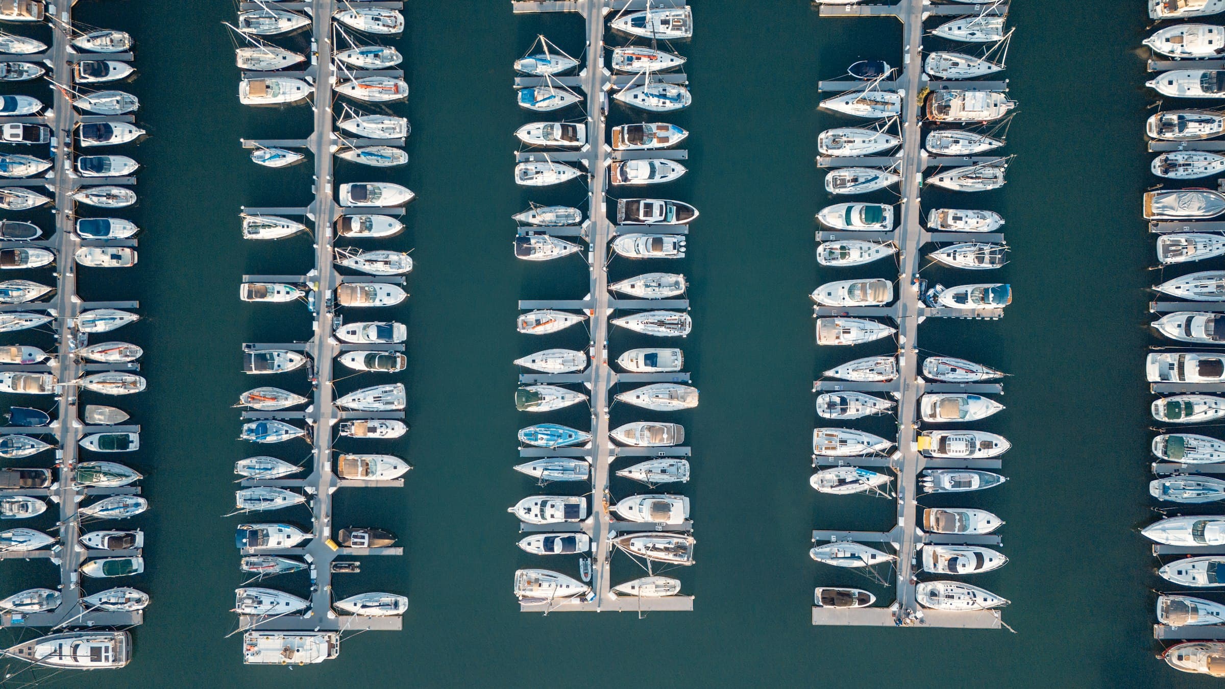 Overhead aerial view of a marina, rows of white vessels berthed along pontoons in dark green water